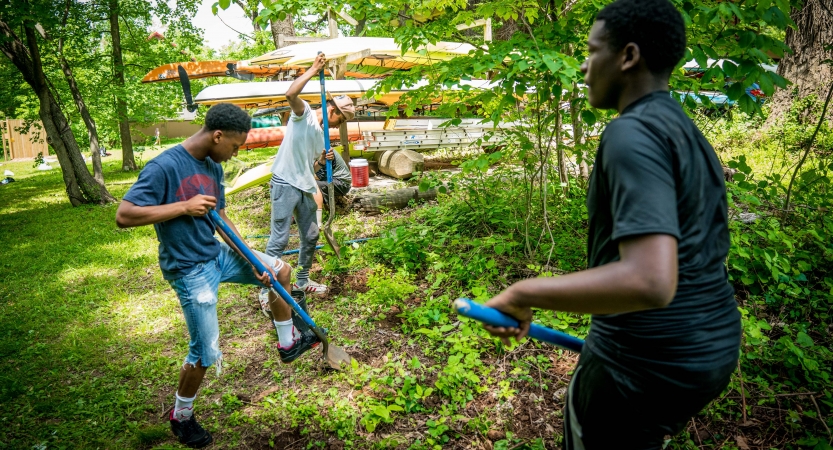 People use gardening tools in a green space. There are canoes on a rack in the background. 